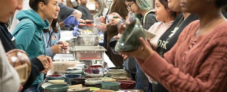 a line of students receive ceramic bowls from across a table of volunteers