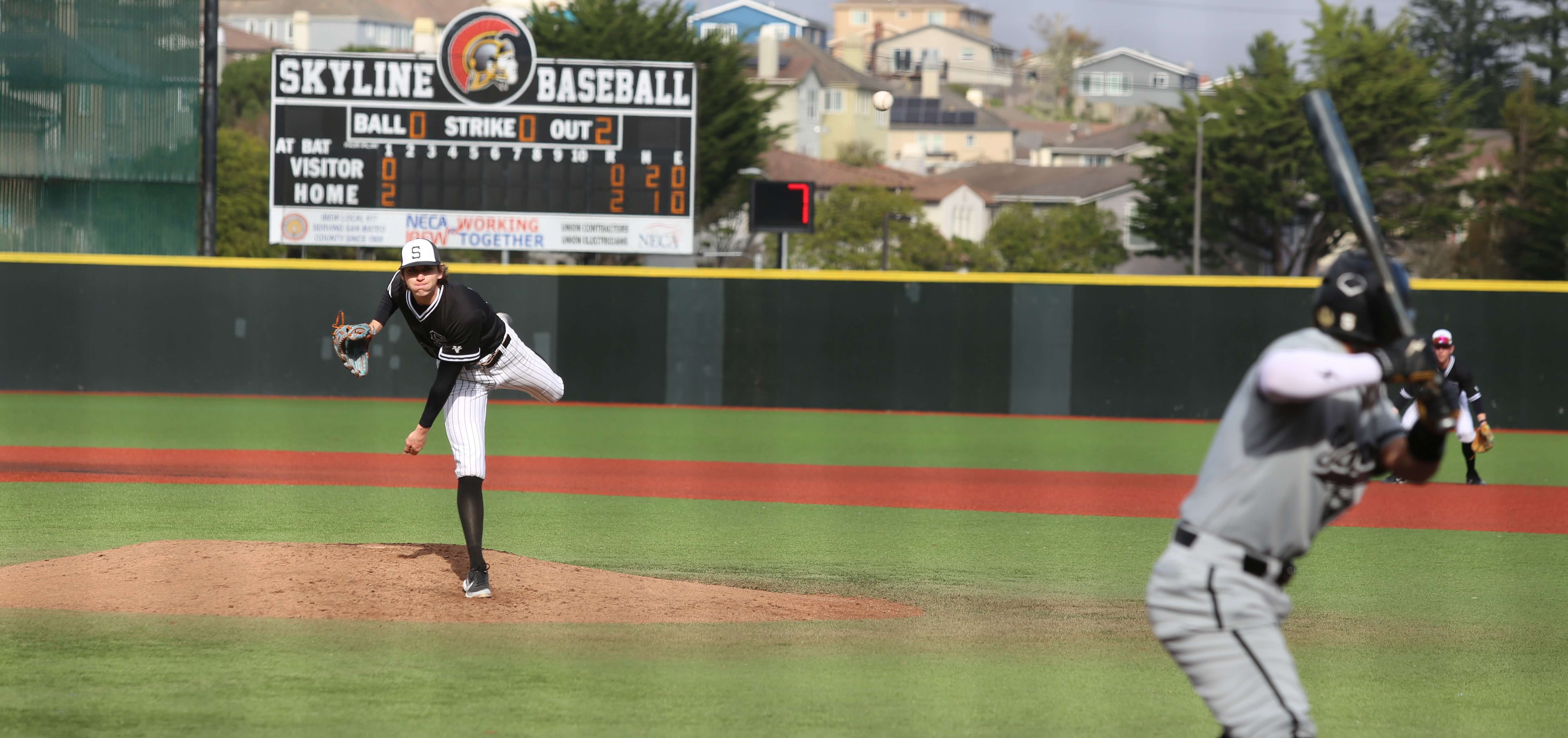 A Skyline College baseball player throws a pitch at an opponent, with the scoreboard in the background showing that Skyline is up by two points.