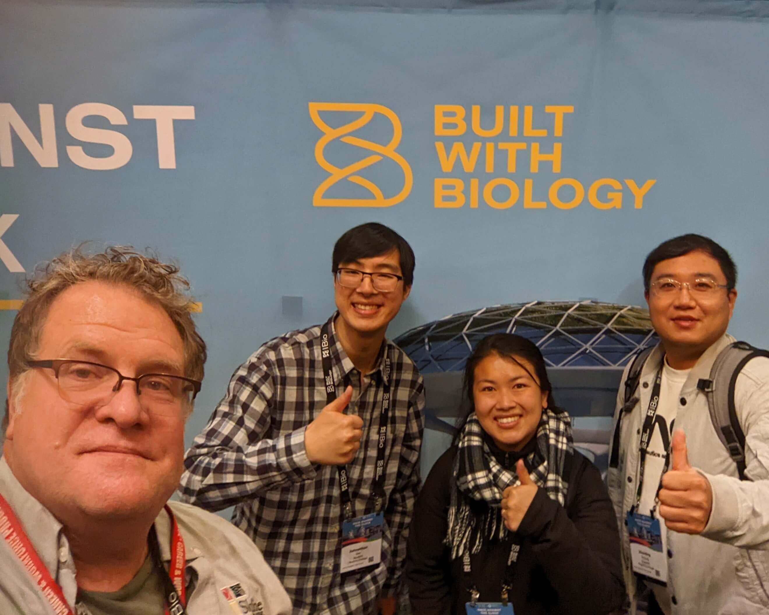 Faculty give thumbs up and stand in front of a backdrop reading 'Built with Biology'
