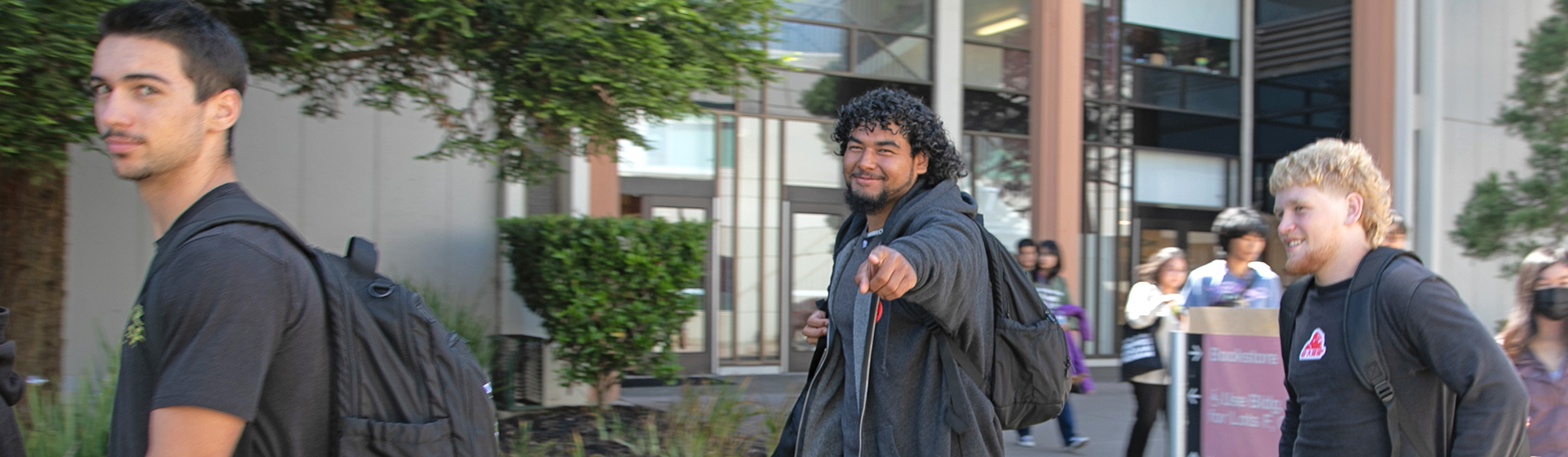 student points at viewer and smiles, walking on campus with fellow students