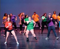 a group of dancers in formation onstage, dressed in shorts and brightly-colored shirts