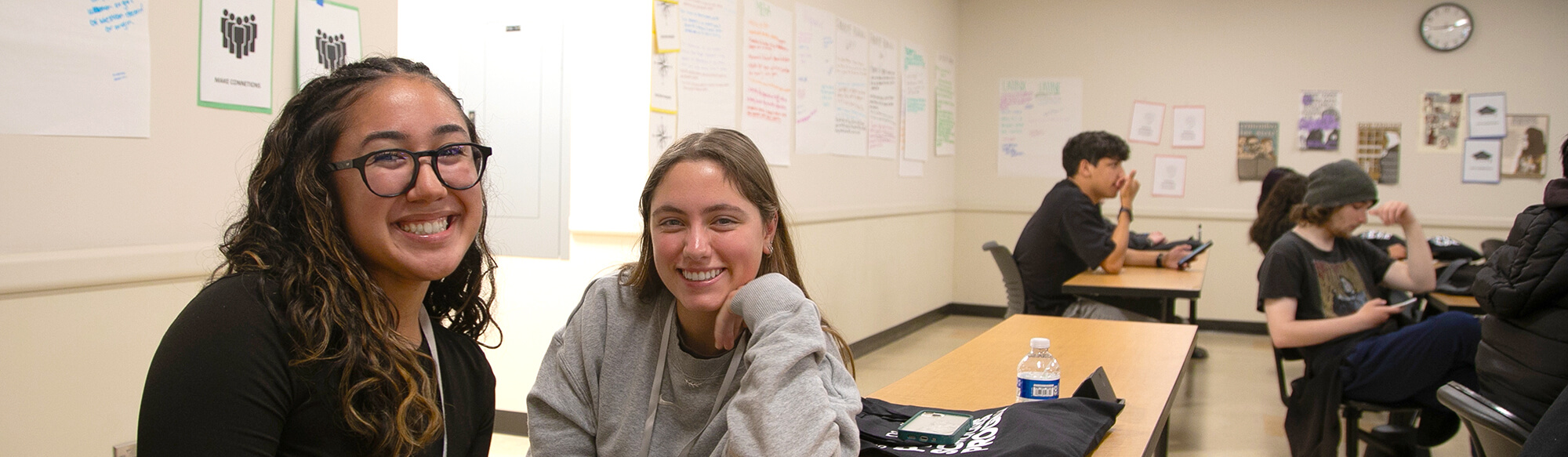 two girls sit at a desk in a classroom and smile