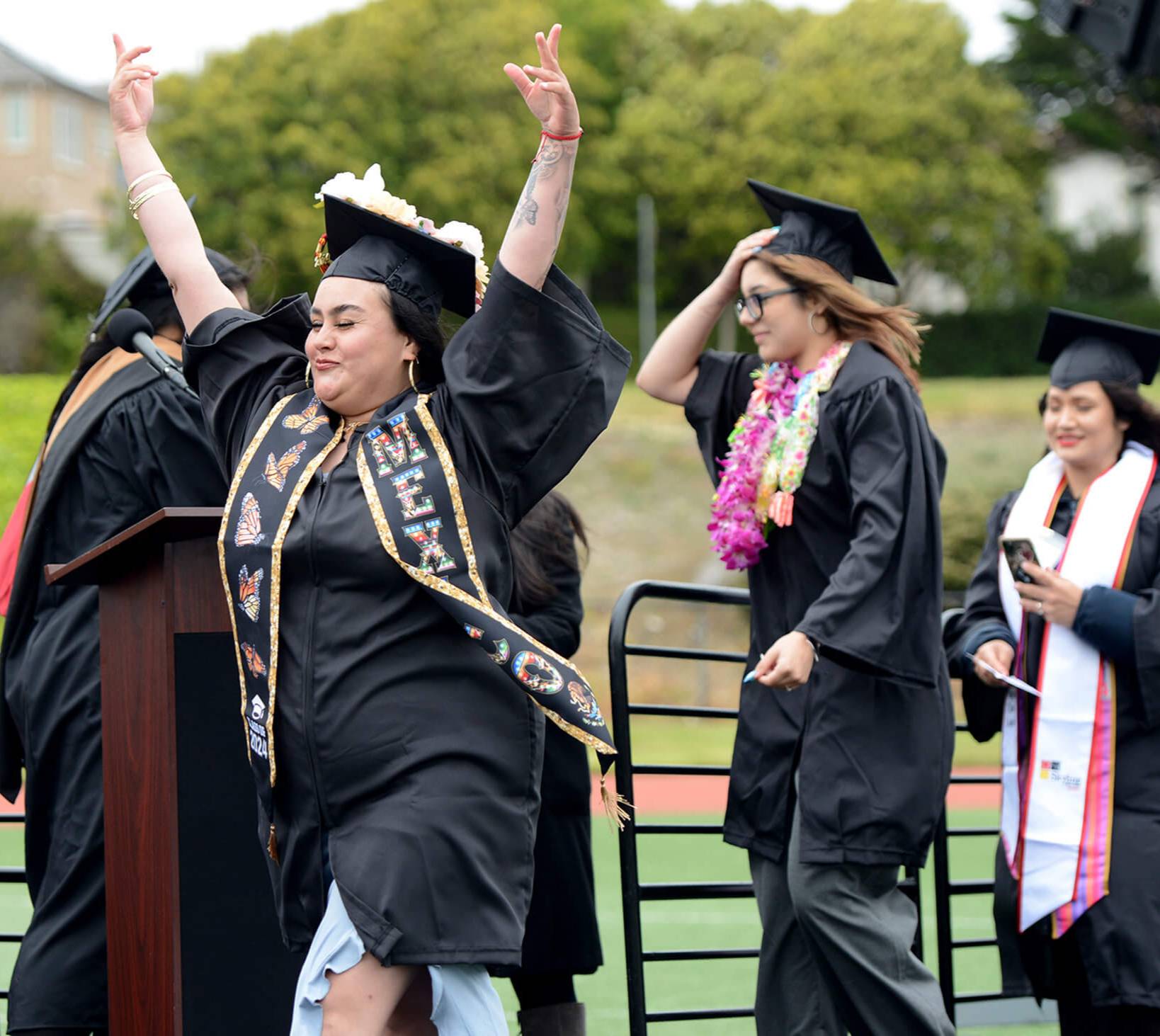 triumphant graduate raises her arms and walks across stage to receive her diploma