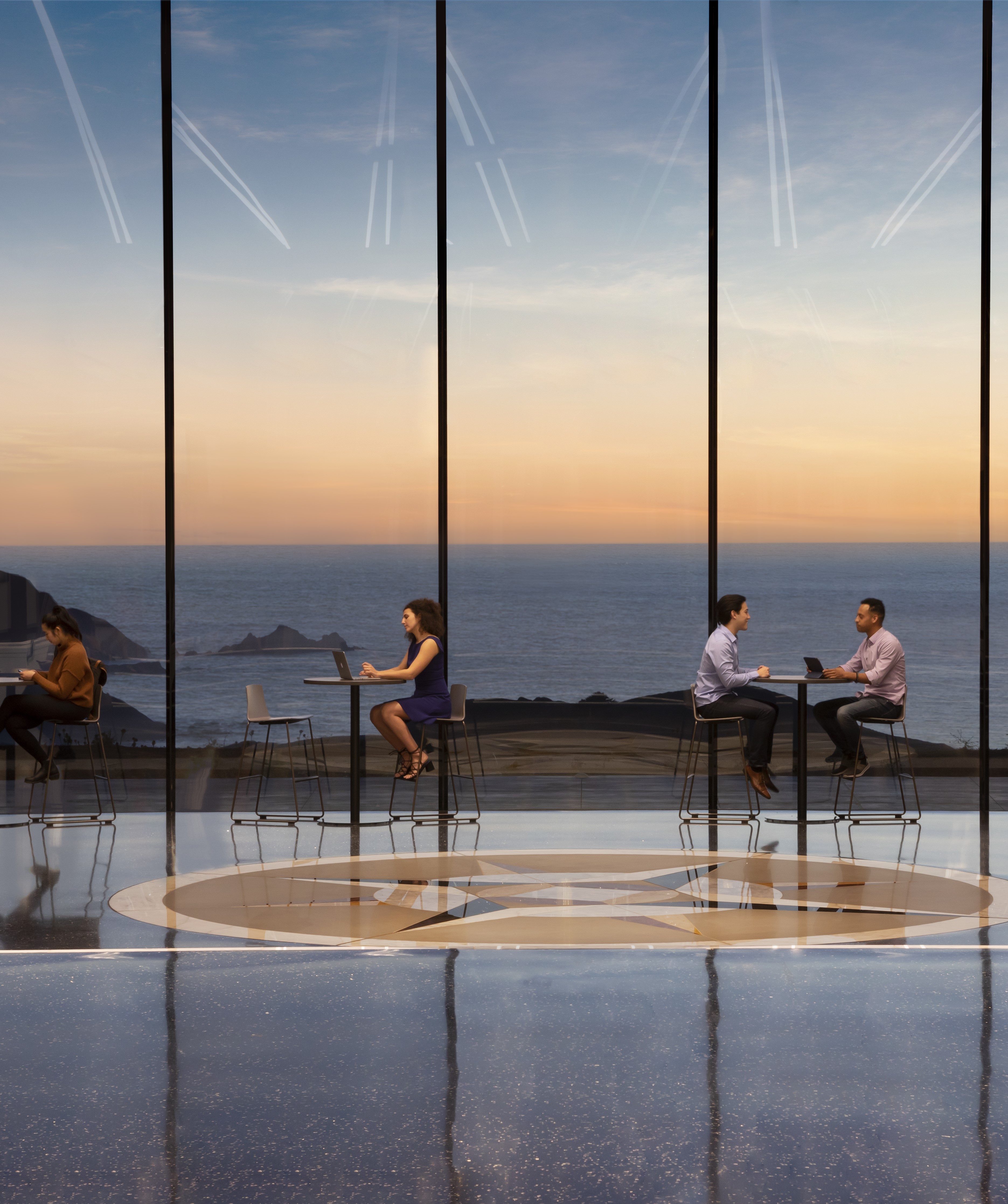 Students study at hightop tables by a floor-to-ceiling window with sunset ocean view inside Building 12
