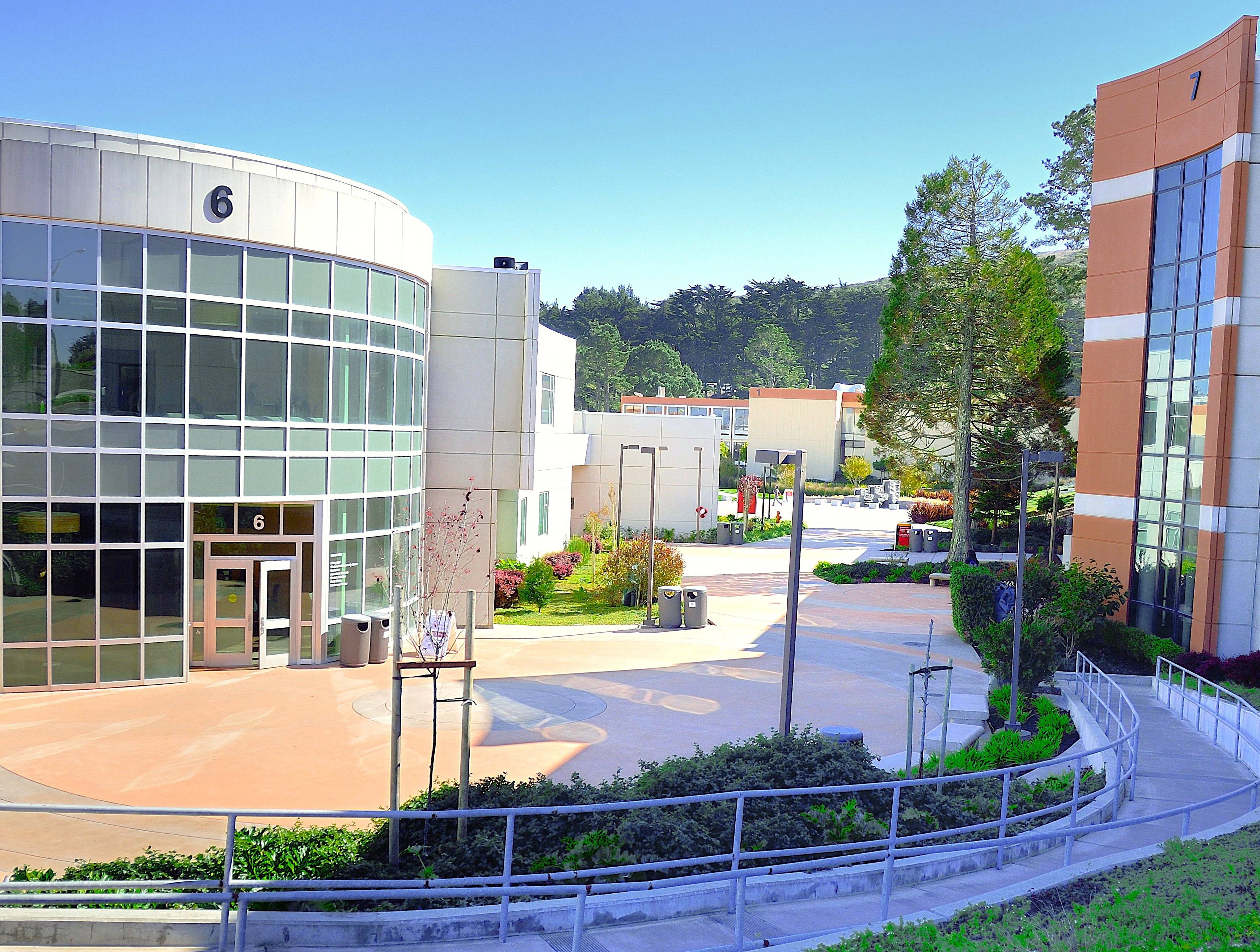 view of the campus route with a ramp leading between the windowed facades of Building 6 and Building 7, with blue skies and greenery