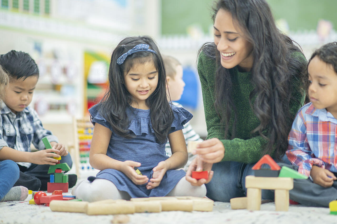 a woman smiles, sitting on the floor and playing with wooden blocks with a group of 4 young children