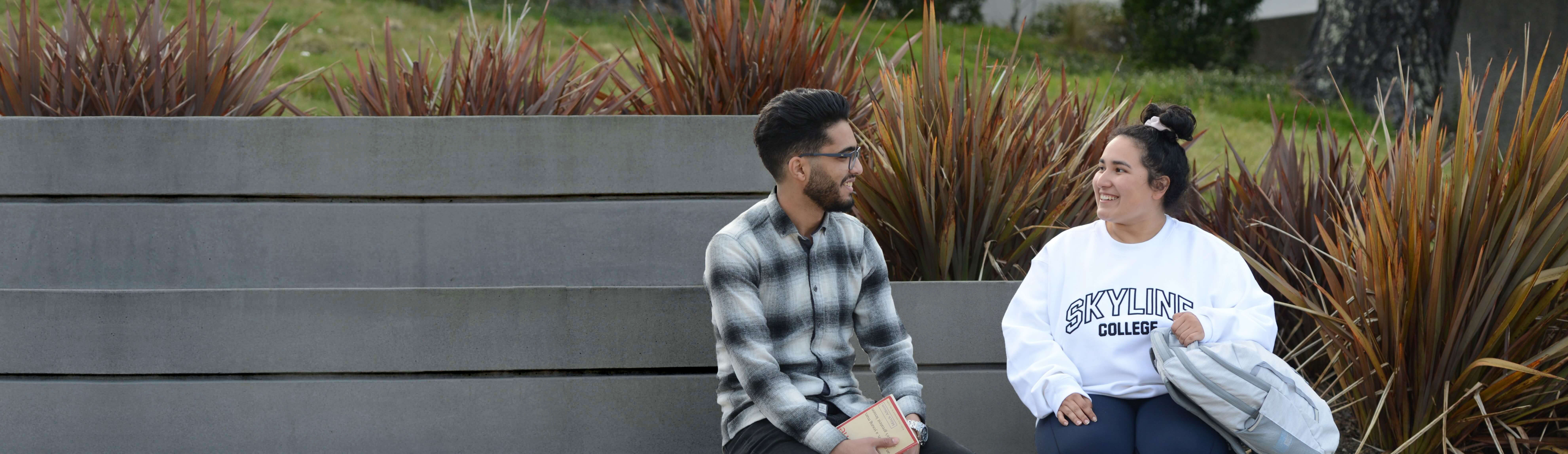 Two students sit together, smiling outside on campus