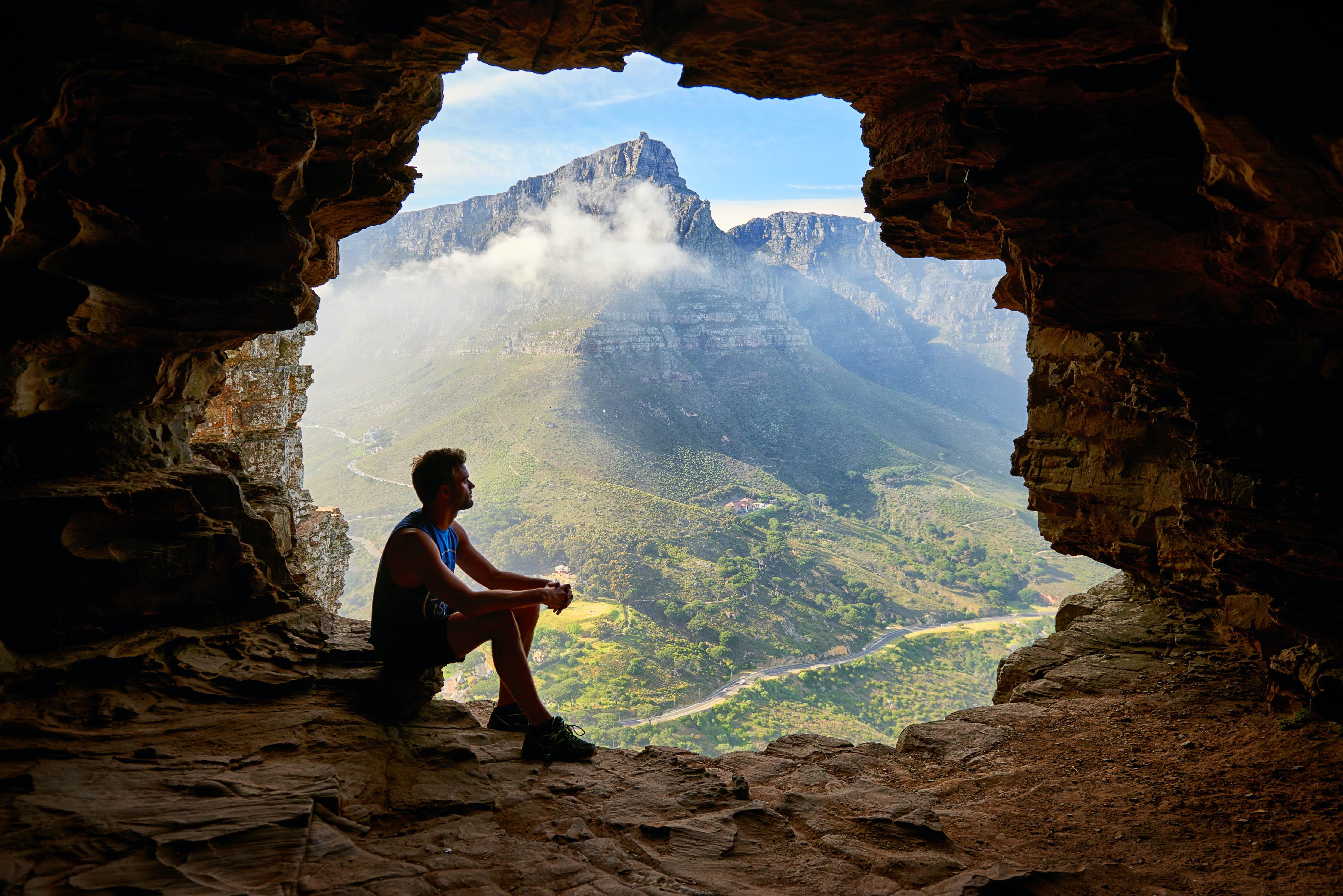 man in hiking gear sits in a natural cave and looks out at a natural monuntain vista